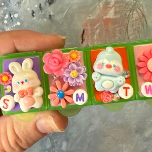 Puede incluir: Un pastillero de pl&aacute;stico verde con cuatro compartimentos, cada uno decorado con un personaje diferente y adornos florales. Los compartimentos est&aacute;n etiquetados con las letras S, M, T y W.