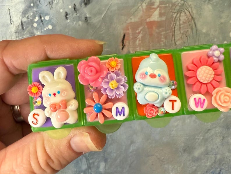 Puede incluir: Un pastillero de pl&aacute;stico verde con cuatro compartimentos, cada uno decorado con un personaje diferente y adornos florales. Los compartimentos est&aacute;n etiquetados con las letras S, M, T y W.