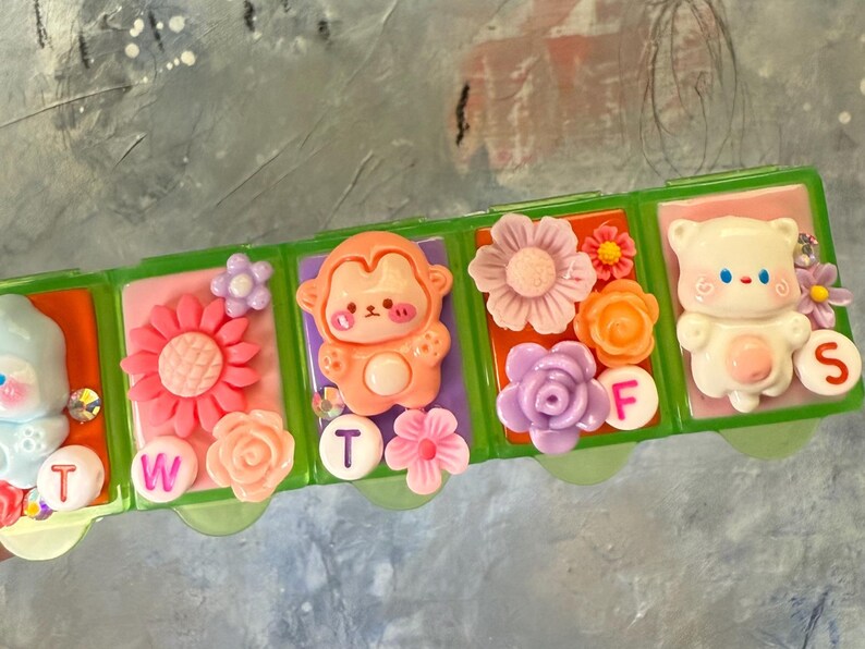 Puede incluir: Un pastillero de pl&aacute;stico verde con compartimentos decorados con personajes de dibujos animados, flores y los d&iacute;as de la semana. Los compartimentos est&aacute;n decorados con un oso, un mono y flores. Las letras T, W, F y S son visibles.