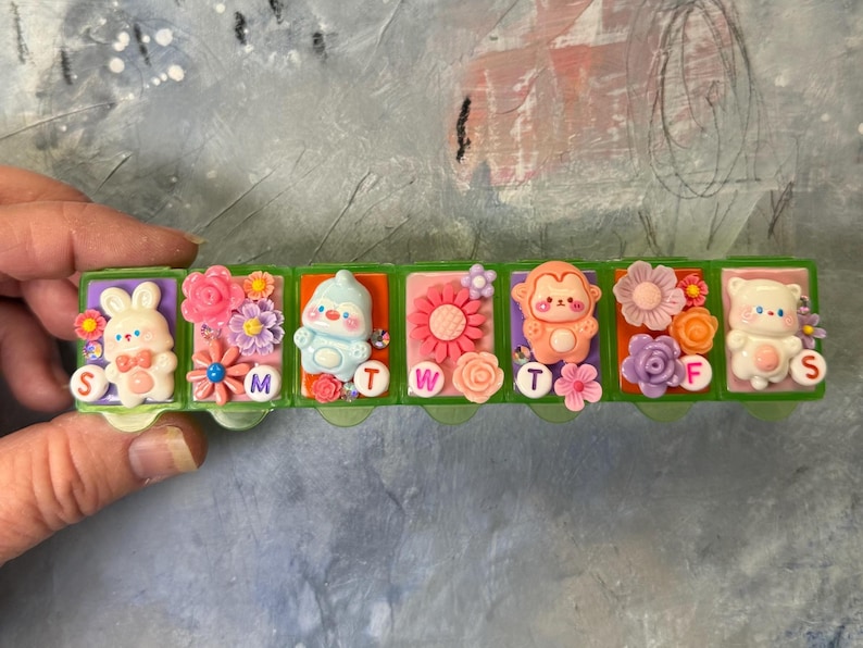 Puede incluir: Un pastillero de pl&aacute;stico verde con forma de tren, decorado con animales de dibujos animados y flores. Cada compartimento est&aacute; etiquetado con un d&iacute;a de la semana. El tren presenta un conejito, un ping&uuml;ino, un mono y un oso, con detalles florales.