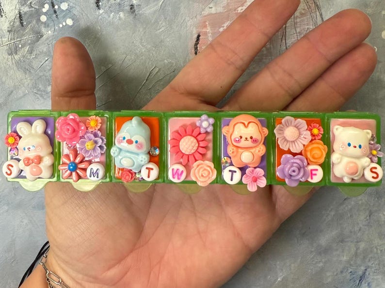 Puede incluir: Un pastillero de pl&aacute;stico verde con cinco compartimentos, cada uno decorado con un personaje de dibujos animados y flores diferentes. Los d&iacute;as de la semana est&aacute;n etiquetados con letras. Los personajes incluyen un conejito, un ping&uuml;ino, un mono y un oso.