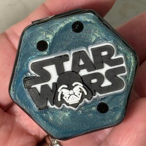 May include: A hexagonal, teal-colored, glittery container with a black and white Star Wars logo and Darth Vader's face in the center.
