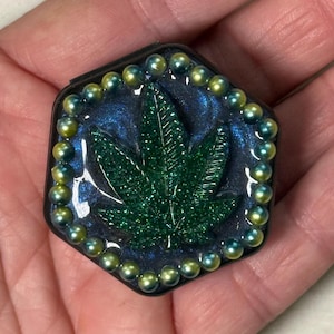 May include: A hexagonal resin trinket box featuring a green glitter marijuana leaf design. The box is bordered by iridescent green and gold beads.