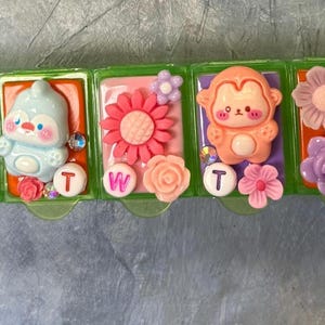 Puede incluir: Un pastillero semanal con dise&ntilde;os decorativos y coloridos. Cada compartimento presenta un dise&ntilde;o diferente, incluyendo un ping&uuml;ino, un mono y varias flores. Los d&iacute;as de la semana se indican con letras en c&iacute;rculos blancos.