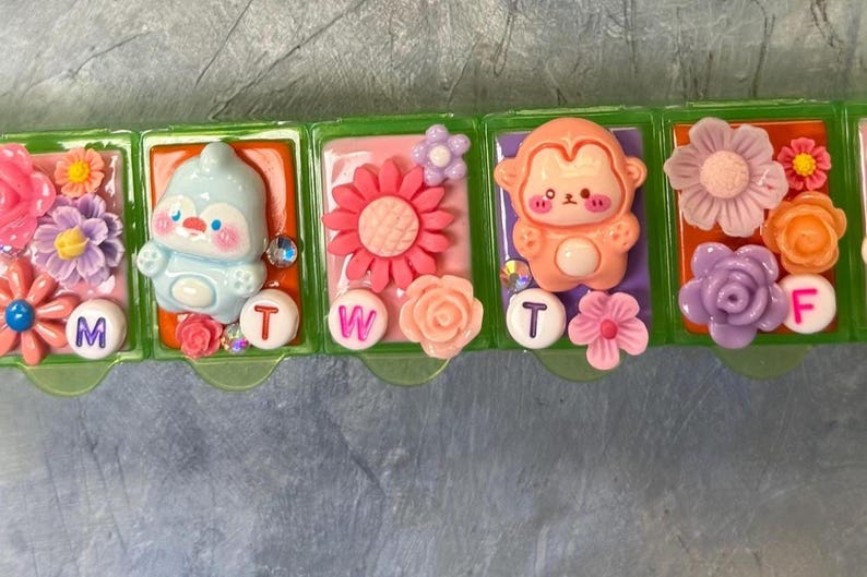 Puede incluir: Un pastillero semanal con dise&ntilde;os decorativos y coloridos. Cada compartimento presenta un dise&ntilde;o diferente, incluyendo un ping&uuml;ino, un mono y varias flores. Los d&iacute;as de la semana se indican con letras en c&iacute;rculos blancos.