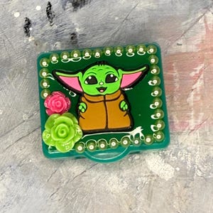 May include: A green square case featuring a cartoon character with large ears, green skin, and a brown coat. The case is decorated with pink and green floral accents and a border of small, white beads.