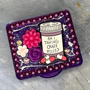 May include: A purple, square trinket box with a decorative border of purple beads. The box is embellished with a pink flower, a white flower, and a purple rose. The box has the text "AM I TAKING CRAZY PILLS?" and an illustration of a pill bottle and pills.