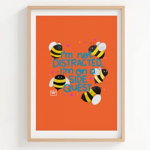 May include: A framed art print featuring an orange background with five cartoon bees and the blue text "I'm not DISTRACTED, I'm on a SIDE QUEST". The bees have yellow and black striped bodies, white wings, and small black antennae. A small white logo is visible.
