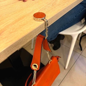 Può includere: Un gancio per borsa argentato con una parte superiore in pelle arancione è attaccato a un tavolo di legno. Una borsa arancione è appesa al gancio. La borsa ha una catena e un cinturino dorati.