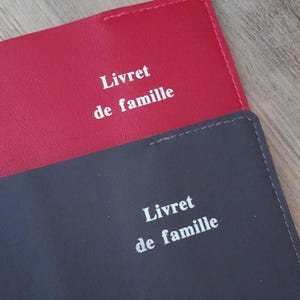 Puede incluir: Se ven dos cubiertas de cuero sintético, una roja y otra gris oscuro. El texto "Livret de famille" está impreso en blanco en ambas cubiertas. Las cubiertas están cosidas con hilo blanco.