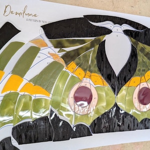 Puede incluir: Un patr&oacute;n de vidriera Galle Moth Demilune con piezas de vidrio numeradas en tonos negro, verde oliva, amarillo brillante y morado oscuro. El dise&ntilde;o incluye una silueta de polilla y una hoja de roble verde sobre una plantilla de papel.