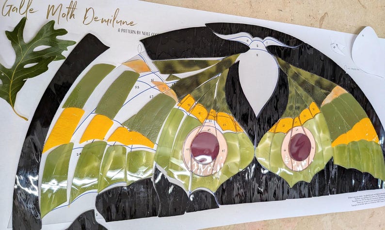 Puede incluir: Un patr&oacute;n de vidriera Galle Moth Demilune con piezas de vidrio numeradas en tonos negro, verde oliva, amarillo brillante y morado oscuro. El dise&ntilde;o incluye una silueta de polilla y una hoja de roble verde sobre una plantilla de papel.