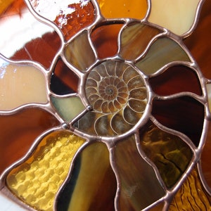 May include: A stained glass art piece depicting a nautilus shell with a spiral pattern. The shell is made of various shades of brown, orange, and yellow glass. The glass pieces are joined together with copper foil.
