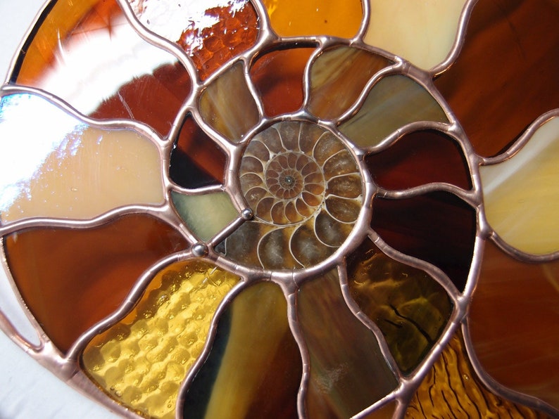 May include: Stained glass art piece in the shape of a nautilus shell. The design features a spiral pattern with various shades of brown, amber, and yellow glass, outlined with copper. The glass has a textured and iridescent appearance.