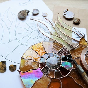 May include: A stained glass ammonite project in progress, featuring a partially assembled piece with iridescent and amber-colored glass. A printed pattern and small ammonite fossils are also visible, along with copper foil and a tool.