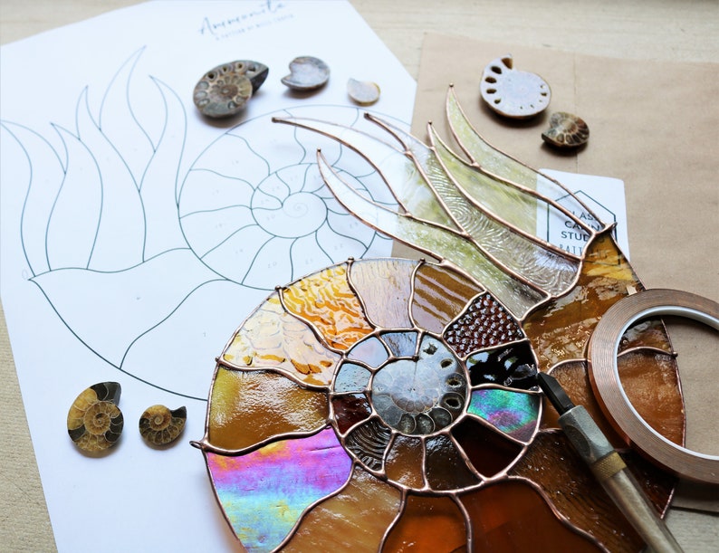 May include: A stained glass ammonite project in progress, featuring a partially assembled piece with iridescent and amber-colored glass. A printed pattern and small ammonite fossils are also visible, along with copper foil and a tool.