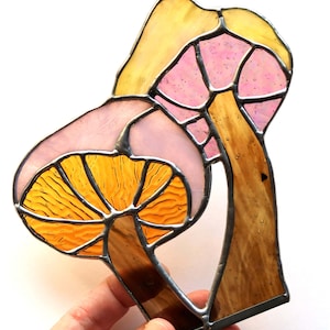 May include: Two stained glass mushrooms, one with a pink cap and the other with a yellow cap, with brown stems. The mushrooms are joined together at the stems.