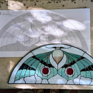 Puede incluir: Un panel de vidrieras semicircular con un dise&ntilde;o de polilla en tonos verde azulado, blanco y negro con acentos circulares rojos. Detr&aacute;s de la pieza de vidrio hay un patr&oacute;n de papel correspondiente etiquetado "Luna Moth Demilune" de Max Glass.