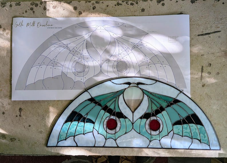 Puede incluir: Un panel de vidrieras semicircular con un dise&ntilde;o de polilla en tonos verde azulado, blanco y negro con acentos circulares rojos. Detr&aacute;s de la pieza de vidrio hay un patr&oacute;n de papel correspondiente etiquetado "Luna Moth Demilune" de Max Glass.