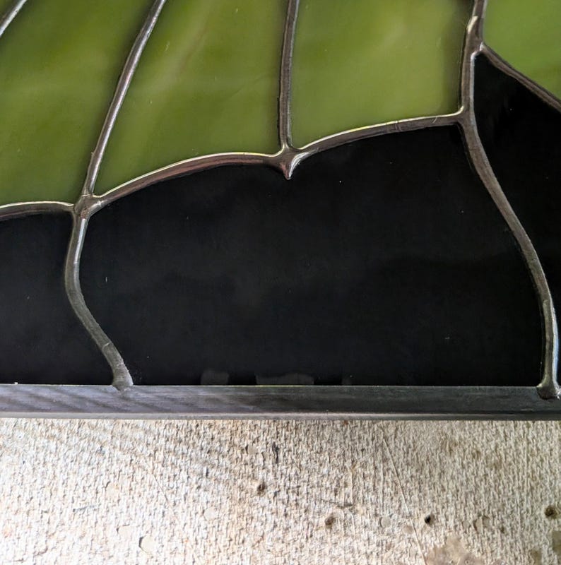 Puede incluir: Un panel de vidriera compuesto por segmentos de vidrio verde oliva transl&uacute;cido y vidrio negro opaco. Las piezas de vidrio individuales est&aacute;n unidas por l&iacute;neas de soldadura de metal oscuro, creando un patr&oacute;n geom&eacute;trico con bordes curvos.