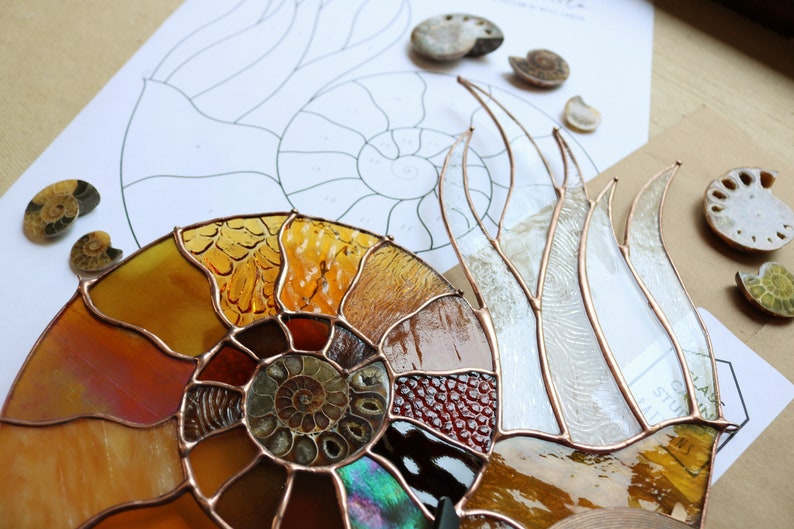 May include: A stained glass ammonite sculpture with copper foil and a variety of glass textures and colors, including amber, brown, and iridescent. The artwork is displayed with fossil specimens and a design sketch.