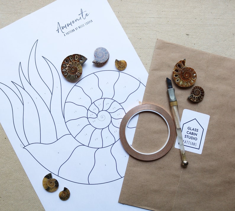 May include: A craft project setup featuring an "Ammonite" pattern by Nellie Cooper. The image includes a printed pattern, several ammonite fossils, a craft knife, copper foil tape, and a Glass Cabin Studio Patterns label.