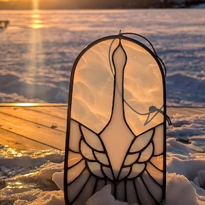 May include: A stained glass window hanging with a white bird design. The bird has a long neck and a wingspan that is wider than the body. The stained glass is set against a snowy background with a wooden deck in the foreground.