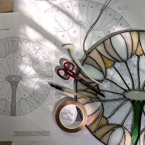 以下が含まれることがあります： 白い表面に置かれた、大きく白とピンクの花のデザインをしたステンドグラスのプロジェクト。花は、パターンシート、ハサミ、テープが近くに置かれた白い表面に置かれています。パターンシートには「Puffball, Pattern by Neill Cooper」と書いてあります。