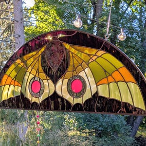 Puede incluir: Un panel de vidriera semicircular con un dise&ntilde;o de polilla en tonos verde lima, naranja y marr&oacute;n oscuro. Las alas de la polilla muestran prominentes acentos circulares de vidrio rosa en el centro, enmarcados por metal oscuro.