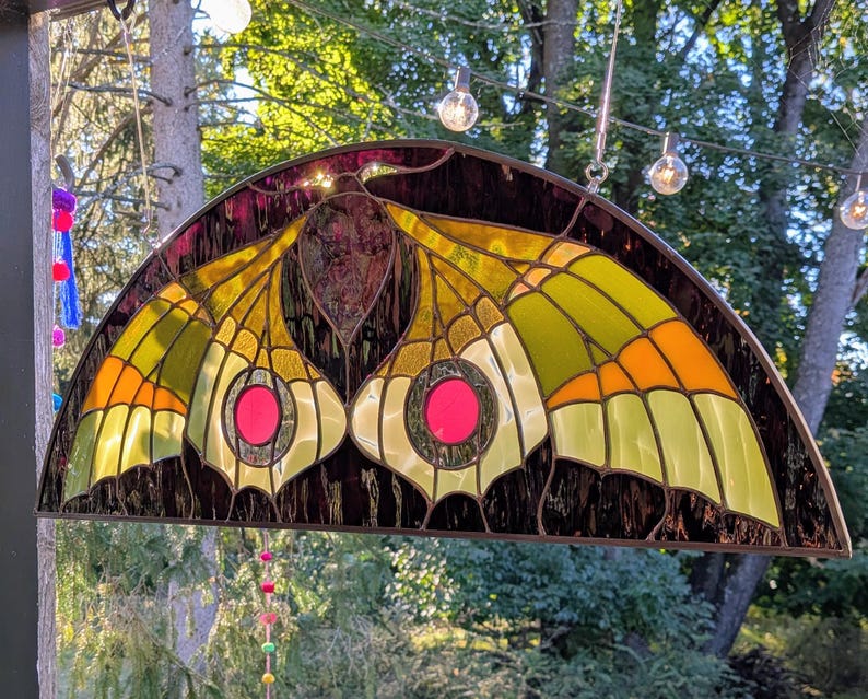 Puede incluir: Un panel de vidriera semicircular con un dise&ntilde;o de polilla en tonos verde lima, naranja y marr&oacute;n oscuro. Las alas de la polilla muestran prominentes acentos circulares de vidrio rosa en el centro, enmarcados por metal oscuro.