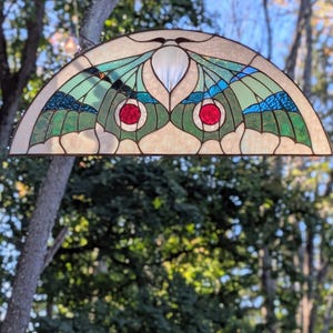 Puede incluir: Un panel de vidriera semicircular con un dise&ntilde;o de polilla, alas de color verde claro, detalles en azul oscuro y dos piezas de vidrio rojo circulares en el centro. El vidrio est&aacute; unido por l&iacute;neas de plomo de metal oscuro.