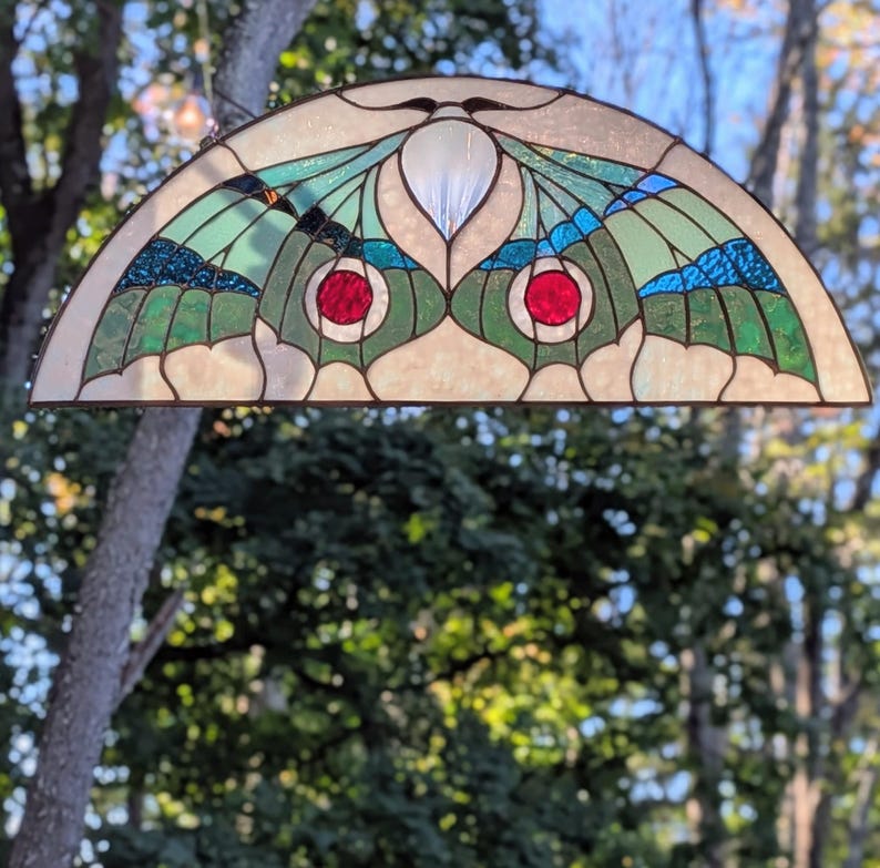 Puede incluir: Un panel de vidriera semicircular con un dise&ntilde;o de polilla, alas de color verde claro, detalles en azul oscuro y dos piezas de vidrio rojo circulares en el centro. El vidrio est&aacute; unido por l&iacute;neas de plomo de metal oscuro.