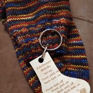 May include: A wooden tag with instructions for the Kitchener stitch, a method for grafting knit fabric together. The tag is attached to a colorful knitted sock with a metal ring.