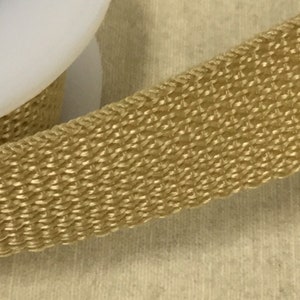 May include: Close-up of a woven, gold-coloured ribbon. The ribbon has a textured surface with a repeating pattern. A white, rounded object is partially visible in the upper left corner. The ribbon is likely used for crafting or decorative purposes.