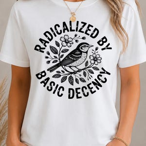 Op de afbeelding: Een wit t-shirt met korte mouwen en een zwarte grafische print van een vogel op een tak, omringd door bloemen. De tekst RADICALIZED BY BASIC DECENCY is in een cirkelvorm rond de centrale vogelillustratie gedrukt.