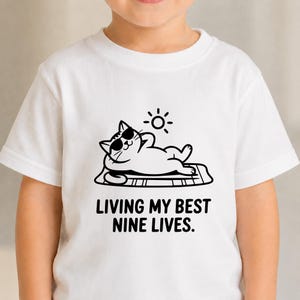 May include: A white short-sleeved t-shirt featuring a black graphic of a cat wearing sunglasses, lying on a towel under a sun. The text below the cat reads, "LIVING MY BEST NINE LIVES." The design is centered on the chest of the garment.