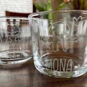 Könnte beinhalten: Zwei klare Glasbecher mit personalisierten Gravuren. Der Becher links hat ein Fuchsmuster und den Namen "Ferd" unten graviert. Der Becher rechts hat ein Krakenmuster und den Namen "Mona" unten graviert.