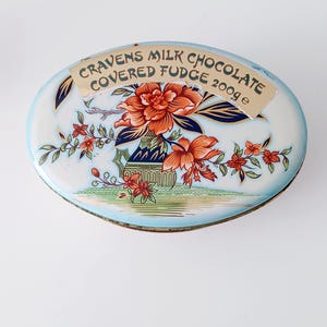 Op de afbeelding: Ovale blikken doos met een bloemenontwerp en de tekst "Cravens Milk Chocolate Covered Fudge 200g e". De doos heeft een lichtblauwe achtergrond met oranje en rode bloemen en groen blad. De doos heeft een lichtblauwe rand.