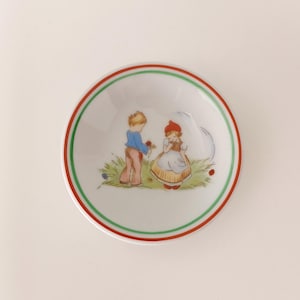 May include: A small, white ceramic bowl with a red and green rim. The bowl features a charming illustration of a boy offering flowers to a girl in a field. The artwork is in soft pastel colours.