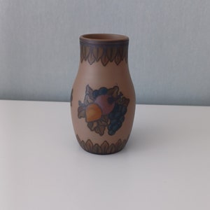Puede incluir: Un pequeño jarrón de cerámica marrón con un diseño pintado de frutas y hojas. El jarrón tiene un cuerpo redondeado y un cuello estrecho.