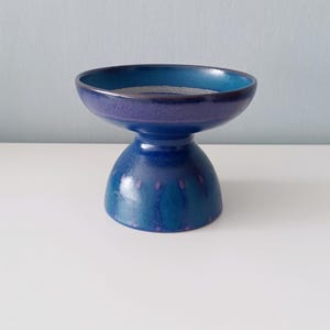 May include: A blue ceramic pedestal bowl with a wide, shallow top and a rounded base. The bowl has a glossy finish with shades of blue and purple. Small, purple dots decorate the base. The bowl sits on a white surface.