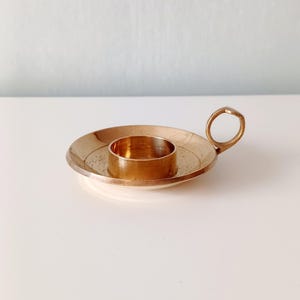 Può includere: Un piccolo portacandele rotondo in ottone con un manico. Il portacandele ha una base circolare e un centro rialzato per contenere una candela. Il manico è un piccolo anello. Il portacandele è su una superficie bianca.
