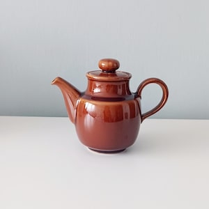 Può includere: Una teiera in ceramica marrone lucido con corpo arrotondato, beccuccio curvo, manico a forma di C e coperchio abbinato con un piccolo pomello arrotondato sulla parte superiore. La teiera presenta una finitura liscia e riflettente di colore uniforme.