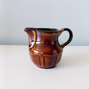 Può includere: Una piccola lattiera in ceramica con una finitura lucida nei toni del marrone. Il contenitore presenta un corpo arrotondato con scanalature verticali modellate alla base, un manico curvo sul lato destro e un beccuccio sul lato sinistro.