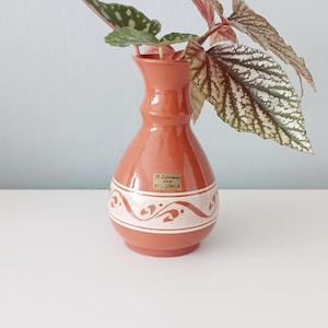 May include: A small, terracotta-colored vase with a white, patterned band around the middle. The vase holds a plant with green and pink leaves. The vase has a label that reads "B. Lehman Holm".