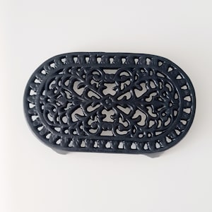 Puede incluir: Un salvamanteles ovalado azul oscuro con un diseño calado ornamentado. El salvamanteles de hierro fundido presenta un detallado patrón floral y de volutas, proporcionando una superficie decorativa y resistente al calor para platos calientes. Mide unos 20 cm de largo.