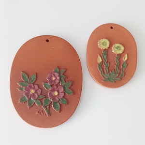 Può includere: Due placche da parete ovali color terracotta. Una presenta un gruppo di fiori viola con foglie verdi, l'altra ha due fiori gialli con steli verdi. Entrambe hanno un piccolo foro per appenderle.