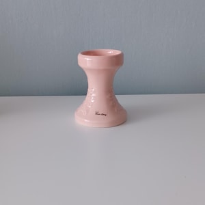 May include: A light pink ceramic candle holder with a flared top and base, connected by a narrower center. The holder features a floral design and the text "Rose Living" on the base. It sits on a white surface.