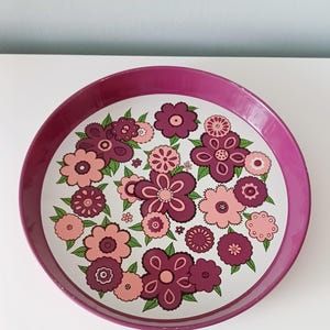 Puede incluir: Bandeja redonda con borde magenta y estampado floral. La superficie de la bandeja presenta un fondo blanco con un patrón repetido de flores estilizadas en tonos rosas y morados, con hojas verdes.