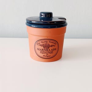 May include: Terracotta marmalade jar with a dark blue lid. The jar has a label that reads "MARMELADE" and "Designed and Crafted in Portugal". The jar is a light orange color and sits on a white surface.